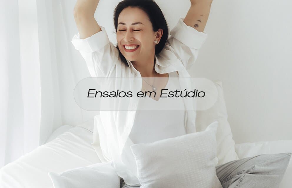 Uma mulher de cabelos curtos está sentada em uma cama de casal com lençóis e travesseiros brancos e macios. Ela está com os braços erguidos e flexionados, as mãos atrás da cabeça, e os olhos fechados, sorrindo abertamente. Ela veste uma camisa branca de botão aberta sobre uma regata branca e calça cinza clara. O fundo é uma janela clara com cortinas brancas e difusas. Uma sobreposição de texto em português no centro diz Ensaios em Estúdio.